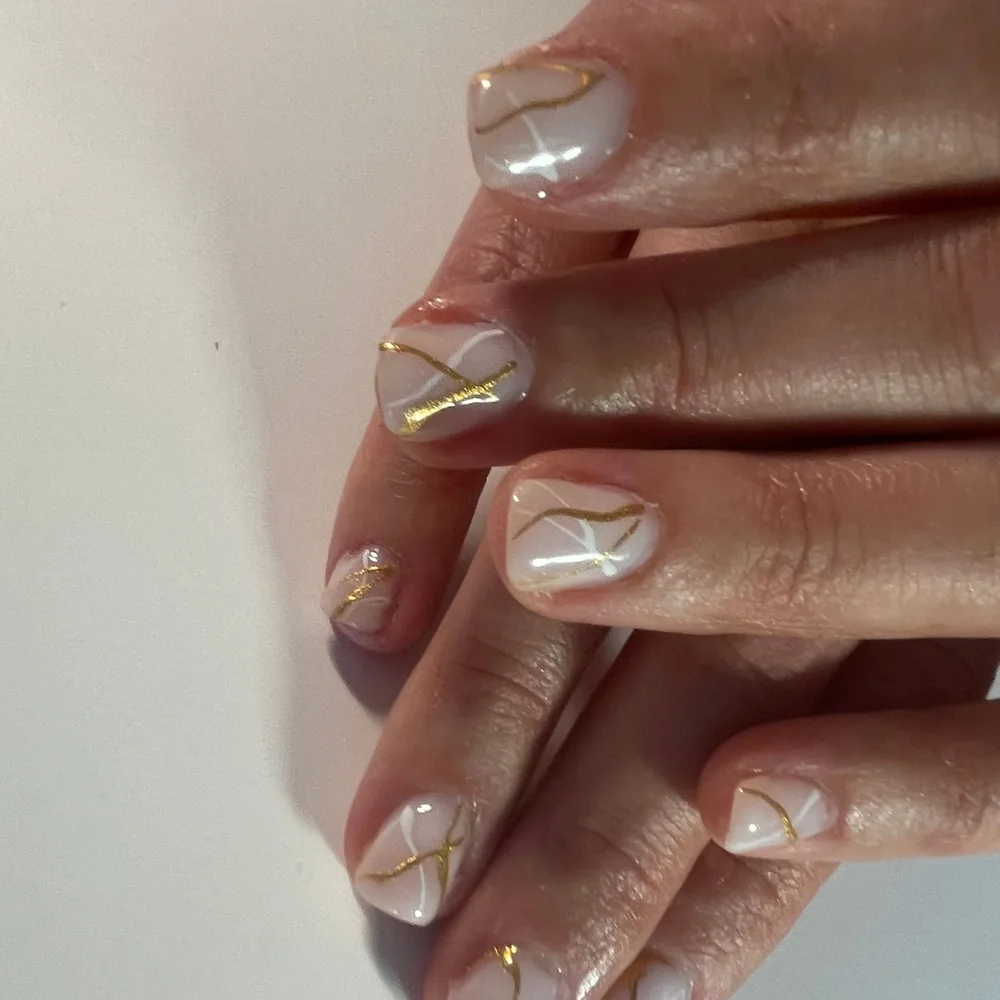 Pose blanc laiteux nacrée sur ongles courts arrondis, filets dorés tracés à main levée